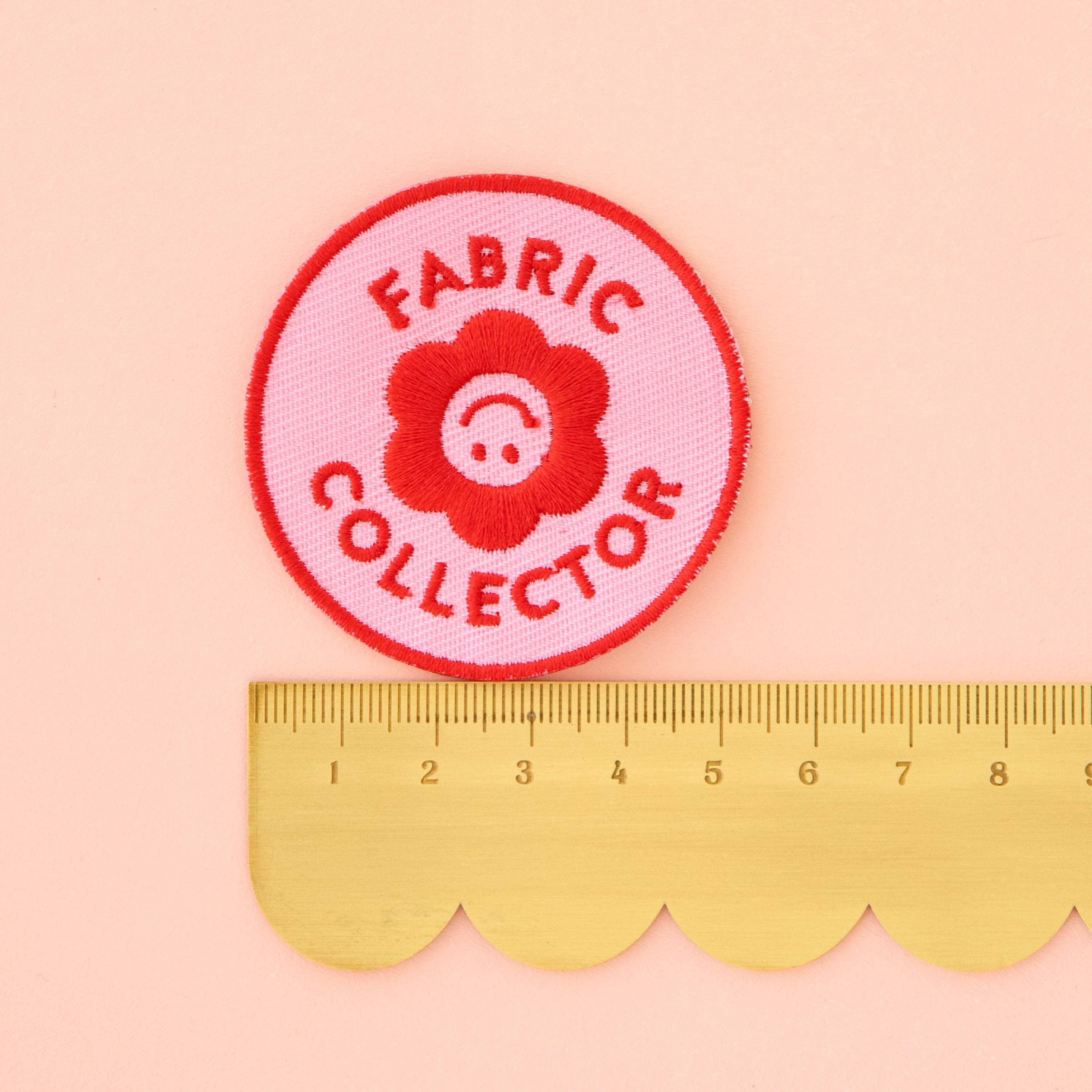 Pink and Red Iron on Patch that says Fabric Collector with a floral smiley face in the middle. Measuring at 5cm.