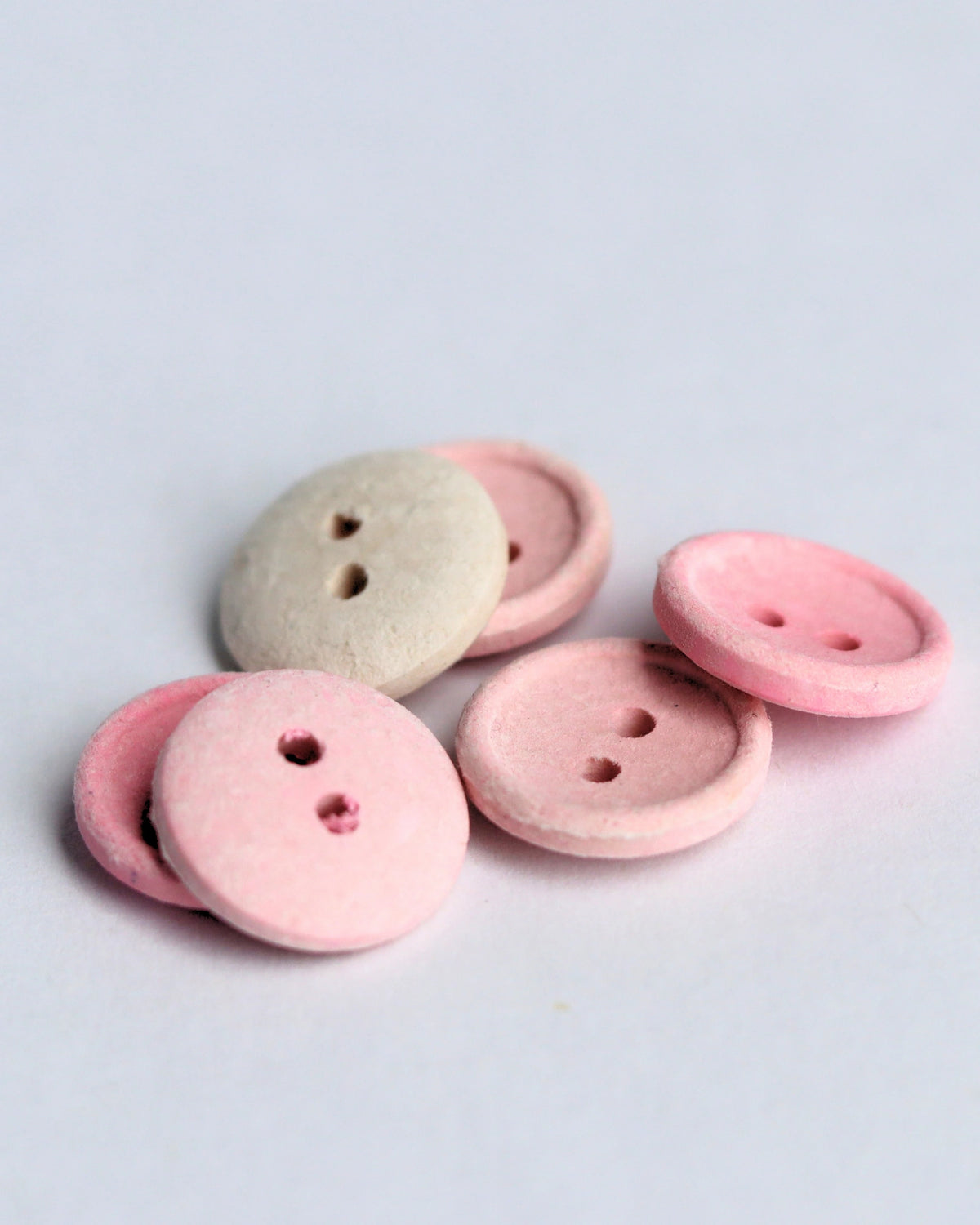 Five small round buttons in soft pastel colors, including pink and a light warm beige, arranged on a plain light gray background.