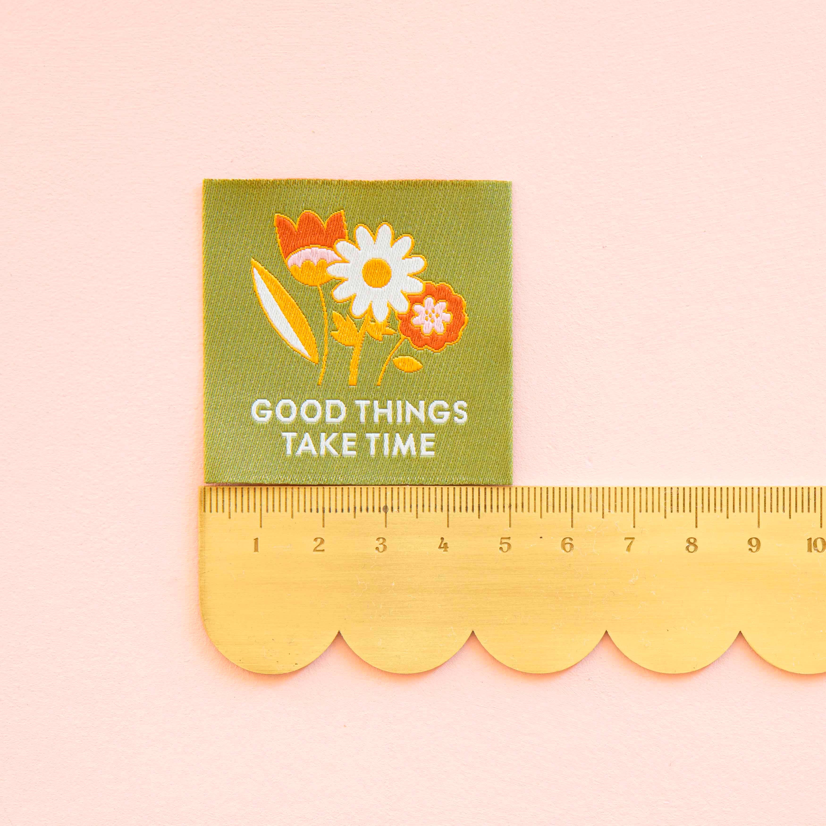 Woven Sew in Label by Sarah Hearts, laid out on a green background is a floral design with the words "Good Things Take Time" a ruler below measures the label at 5cm wide and high.