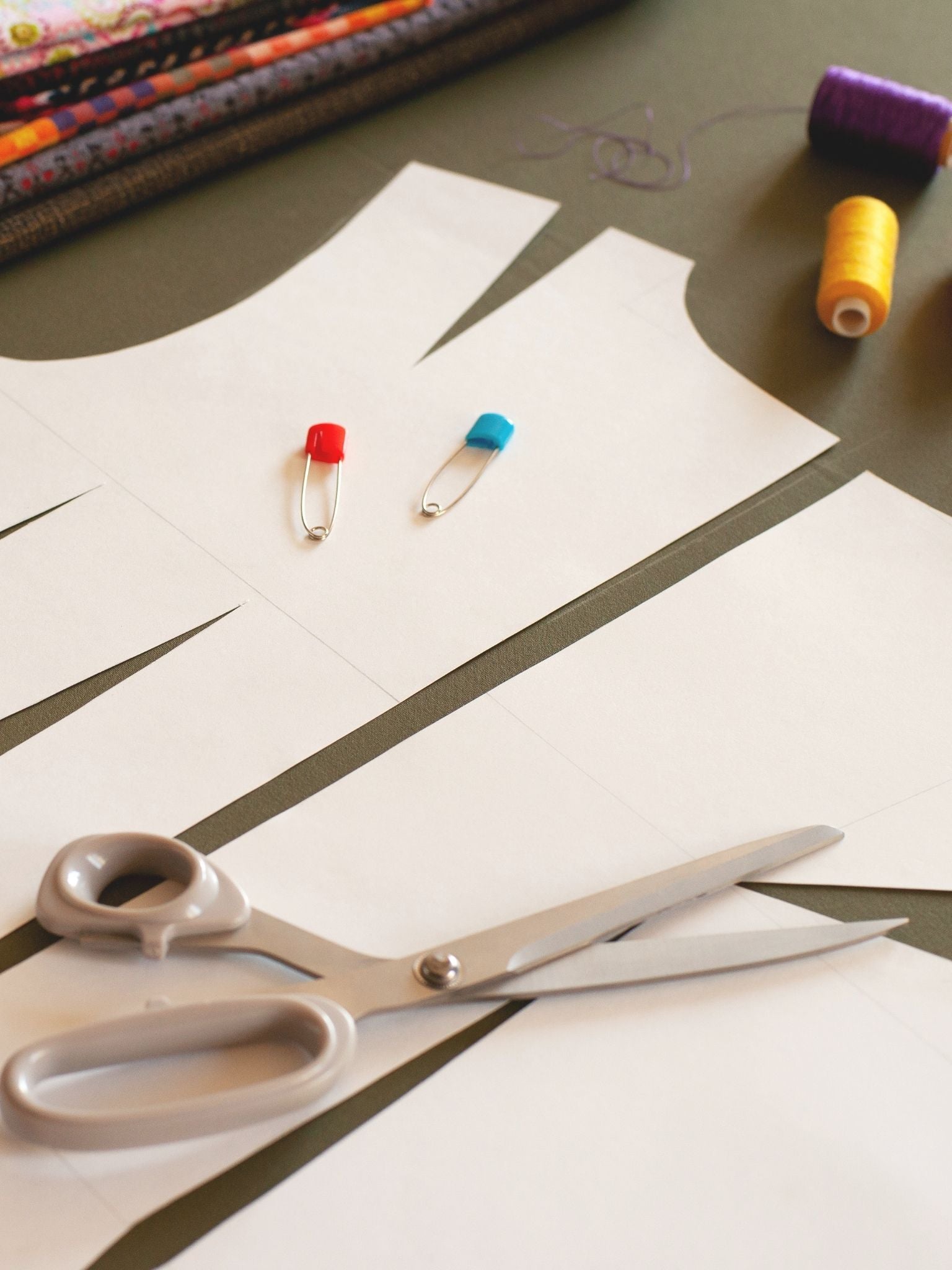 Flat lay of paper sewing pattern pieces with scissors, colourful safety pins, spools of thread, and folded fabrics in the background