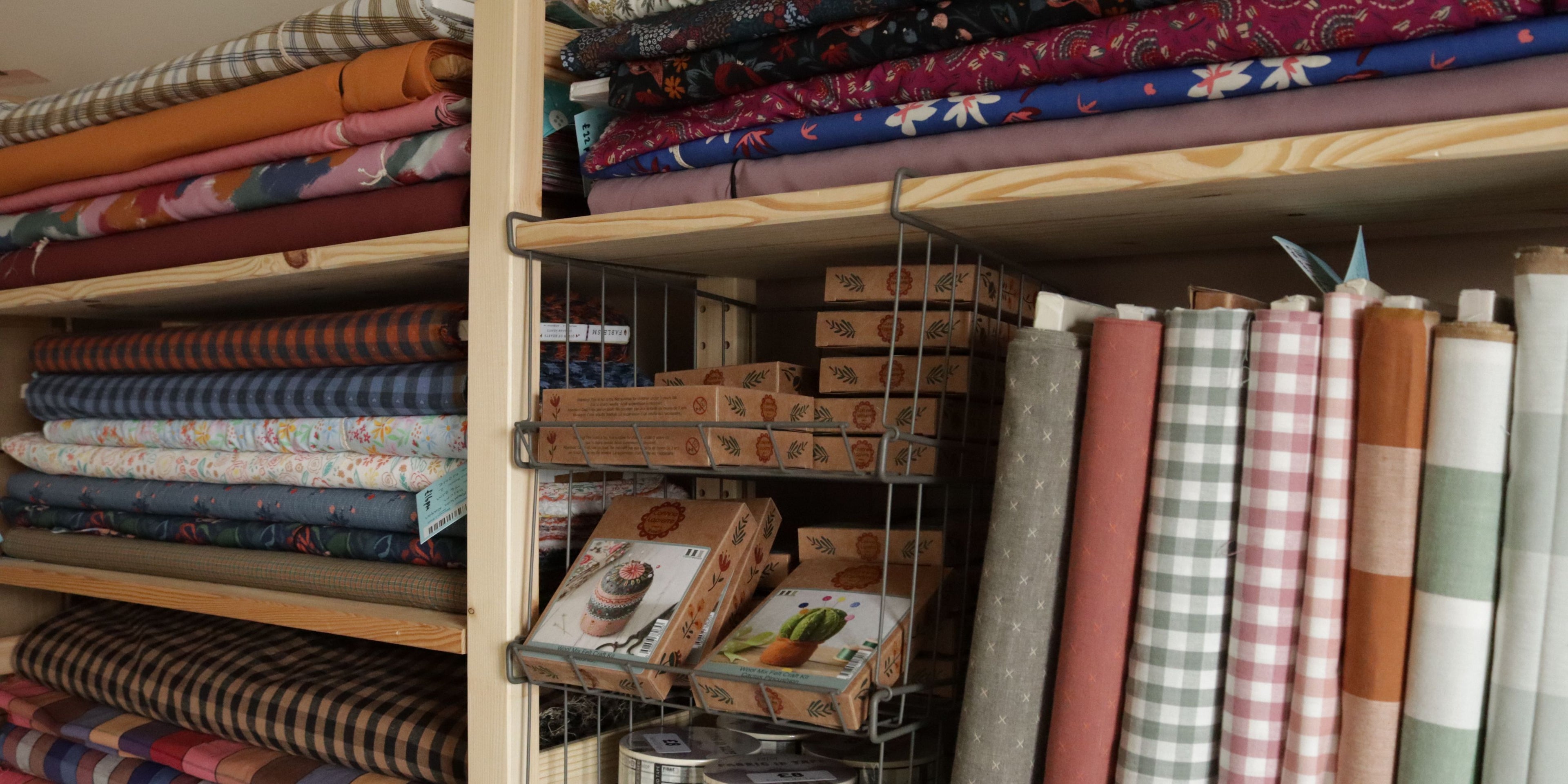 Wooden shelving stacked with folded fabric bolts and haberdashery kits, including gingham, floral, and solid colours, with fat quarters and felt craft kits neatly displayed