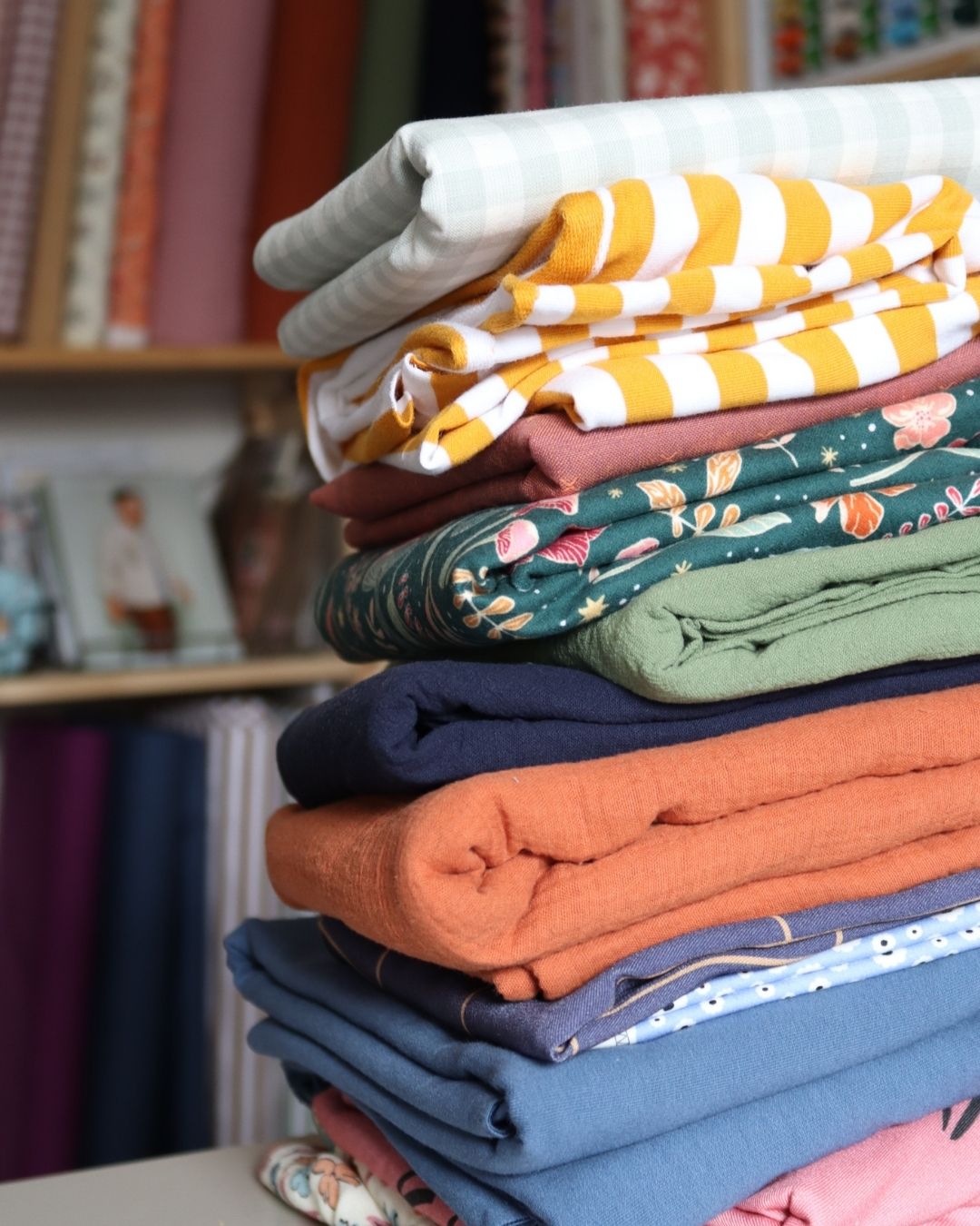 Stack of folded sustainable fabrics in various colours and prints, including stripes, florals, and solids, with sewing supplies in the background