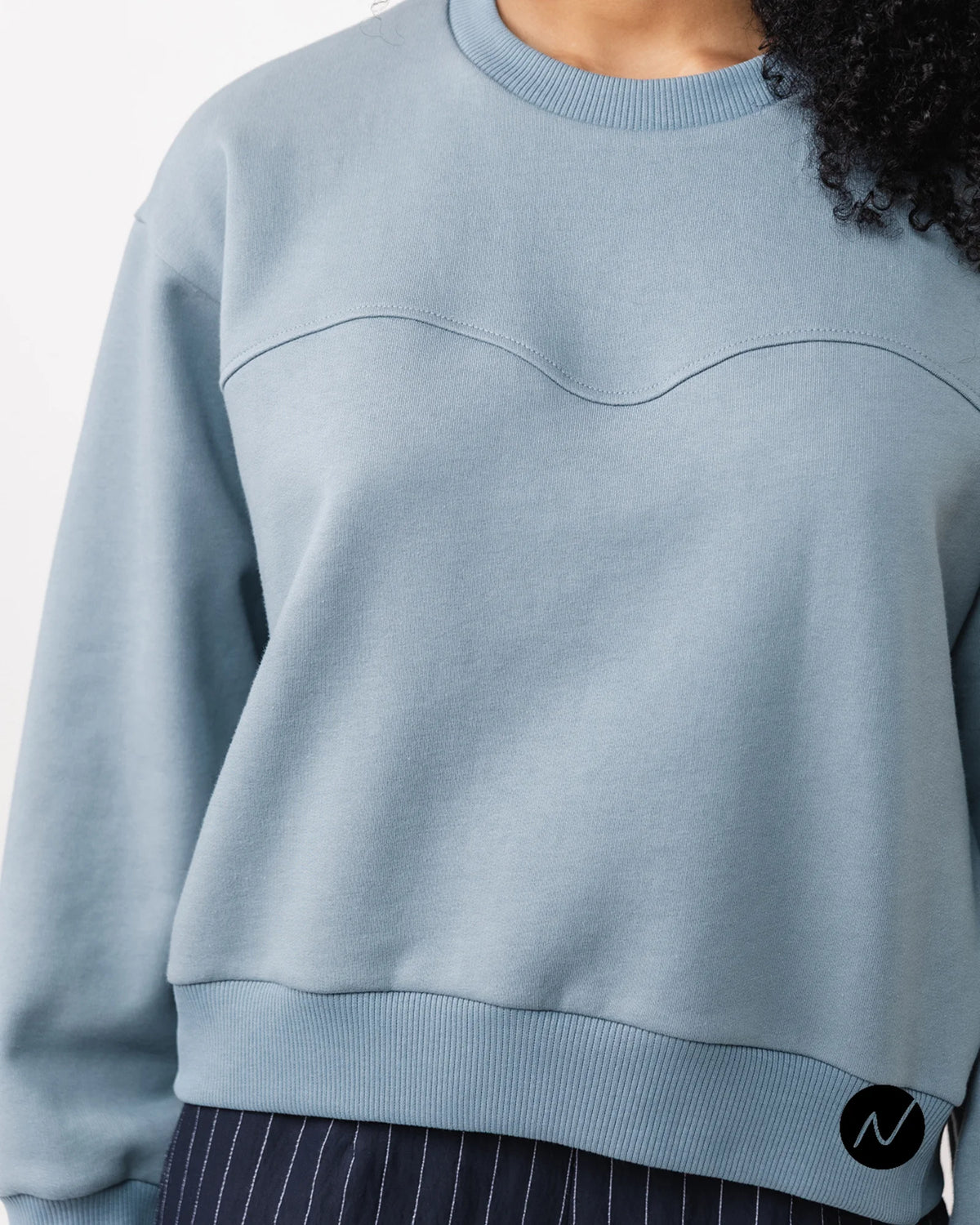 A close-up of a person wearing a light blue crew neck sweatshirt with a wavy chest seam detail, paired with dark pinstriped pants.