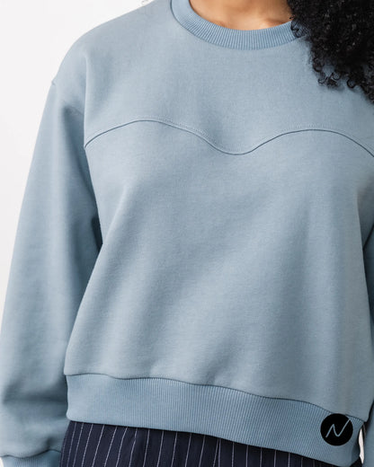 A close-up of a person wearing a light blue crew neck sweatshirt with a wavy chest seam detail, paired with dark pinstriped pants.