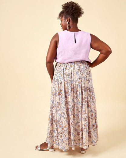 A woman shown from the back wearing a sleeveless lavender top and a long tiered maxi skirt with a beige, brown, and blue botanical pattern, set against a solid beige studio background.