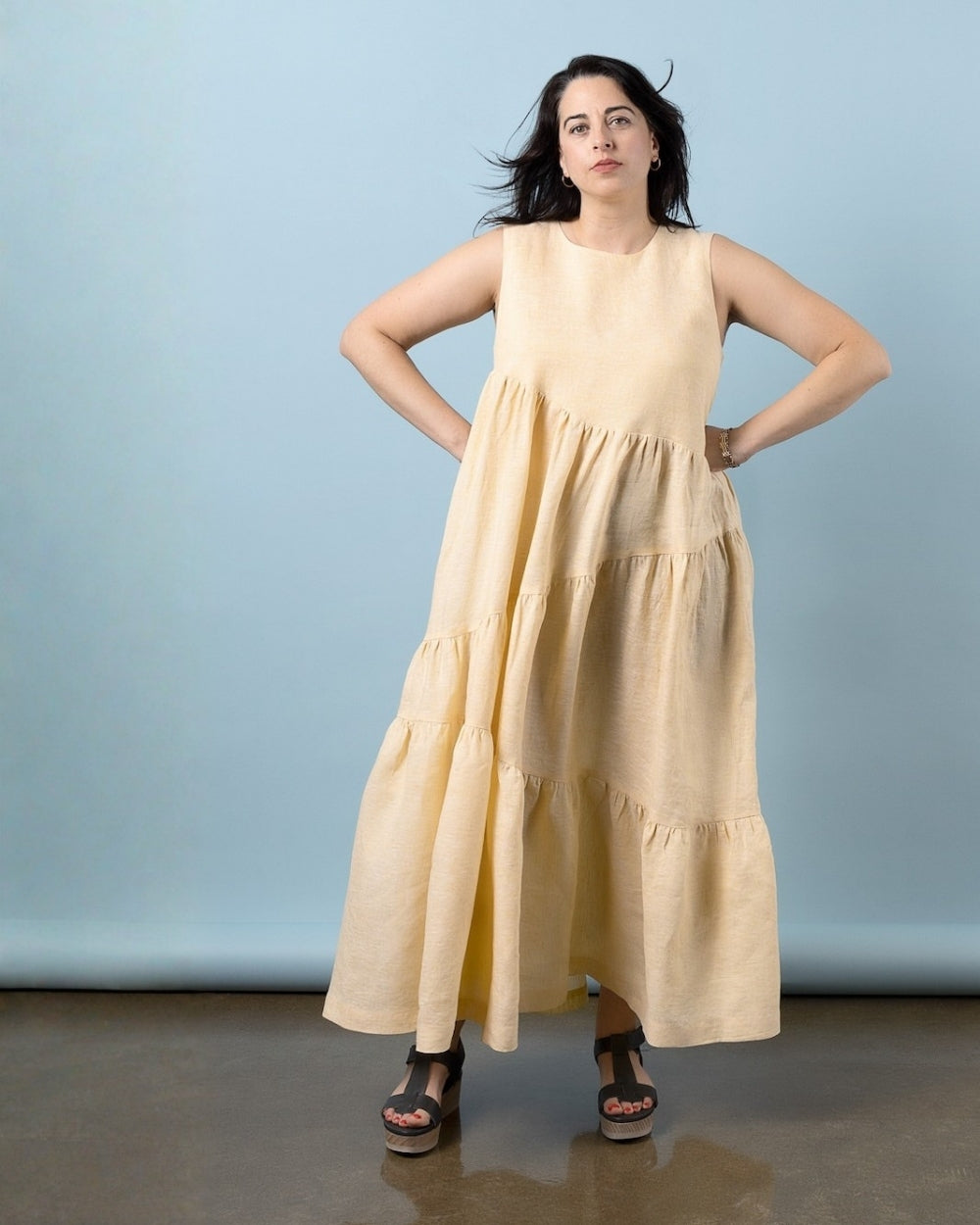 A woman modeling a sleeveless, tiered maxi dress in a light beige or cream color, standing against a solid light blue studio backdrop.