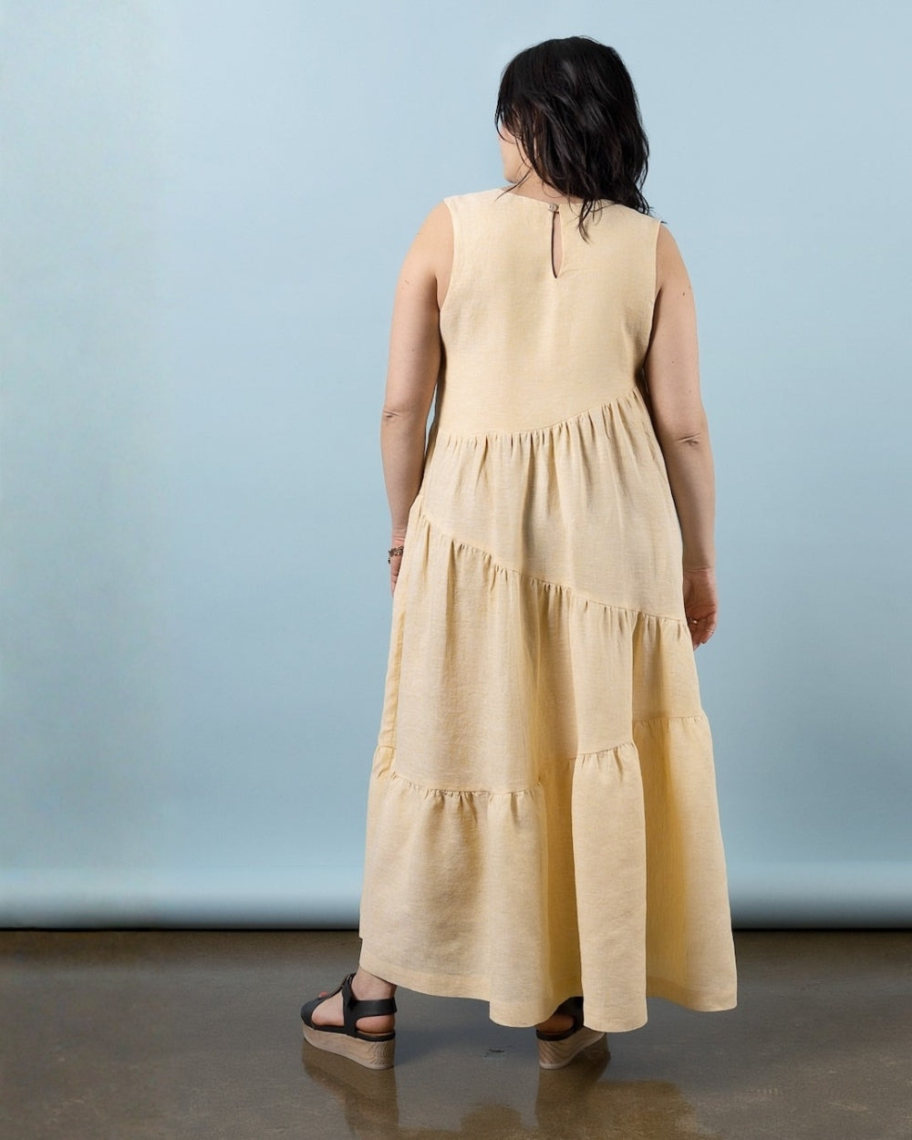 A woman viewed from behind wearing a sleeveless, tiered maxi dress in a light beige or cream color, standing against a light blue studio backdrop.