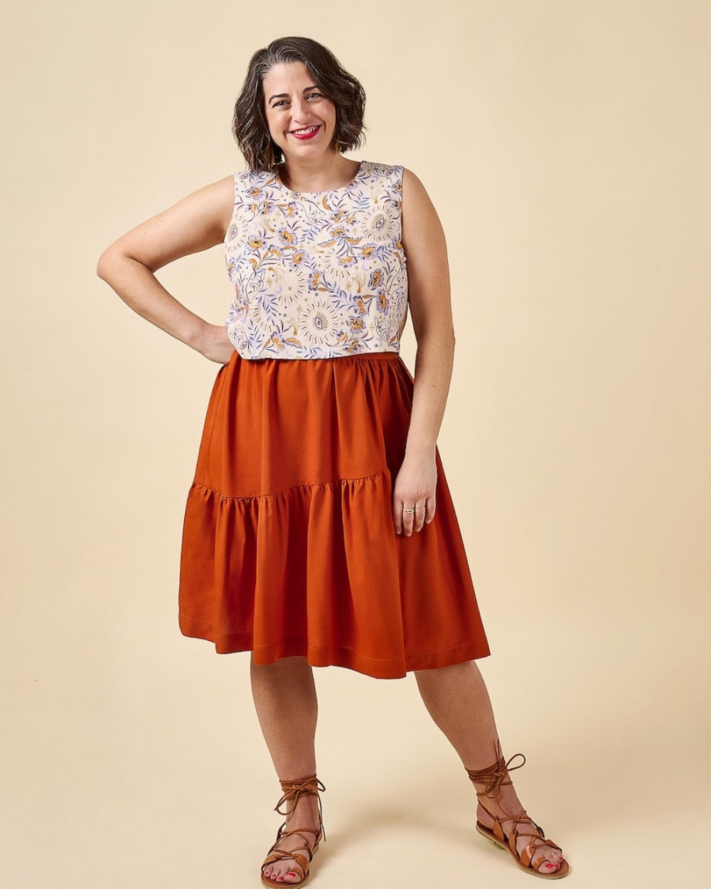 A woman modeling a sleeveless midi dress with a cream floral patterned bodice and a tiered rust-orange skirt, paired with brown lace-up sandals, against a solid beige background.