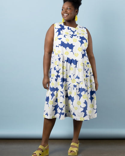 A woman modeling a sleeveless midi dress with a blue, white, and yellow floral pattern, paired with yellow sandals, against a light blue studio background.
