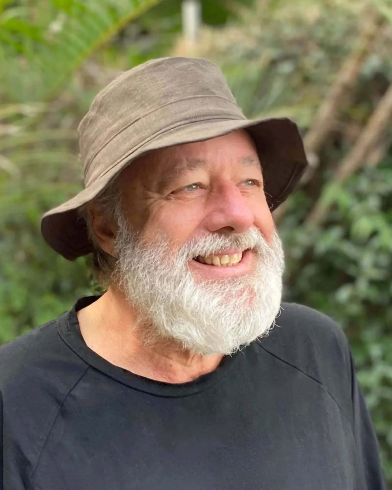 A smiling man with a white beard wearing a brown bucket hat and a black t-shirt, photographed outdoors against a soft-focus green background.