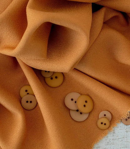 A close-up of draped warm camel-colored fabric with several small decorative buttons in cream, tan, and mustard yellow scattered across the surface.