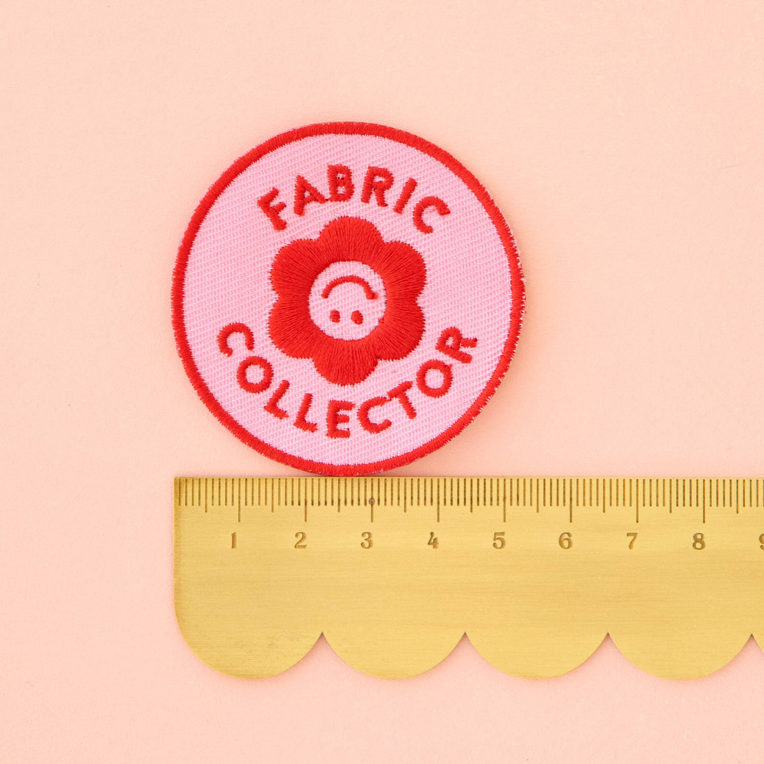 Pink and Red Iron on Patch that says Fabric Collector with a floral smiley face in the middle. Measuring at 5cm.