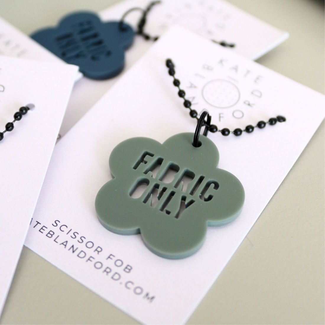 Green fabric-only scissor fob with black beads on a white card with branding.