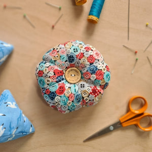 birds eye view of button theme pin cushion, surrounded by pins, threads and embroidery scissors