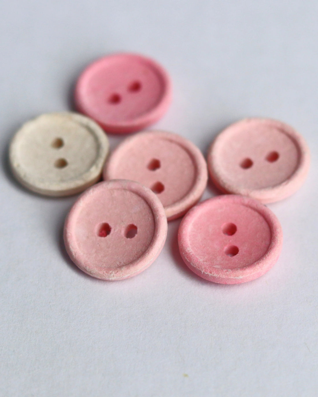 Six small round buttons with two holes, including five shades of pink and one light beige, arranged on a light gray background.