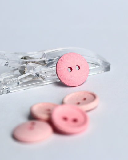 Pink buttons in various sizes, with one larger button resting on a clear plastic clip and four smaller buttons blurred in the foreground on a light gray background.