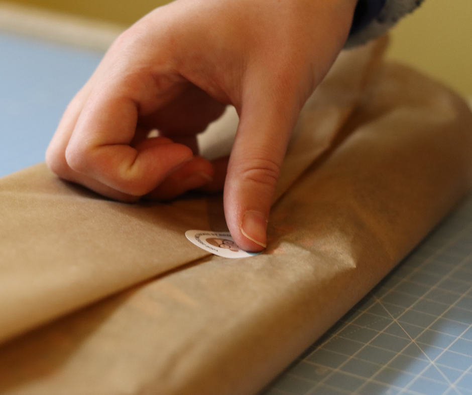 white womans hand, pushing down sticker on kraft tissue paper. Packaging up fabric and sending out orders