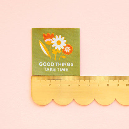 Woven Sew in Label by Sarah Hearts, laid out on a green background is a floral design with the words &quot;Good Things Take Time&quot; a ruler below measures the label at 5cm wide and high.