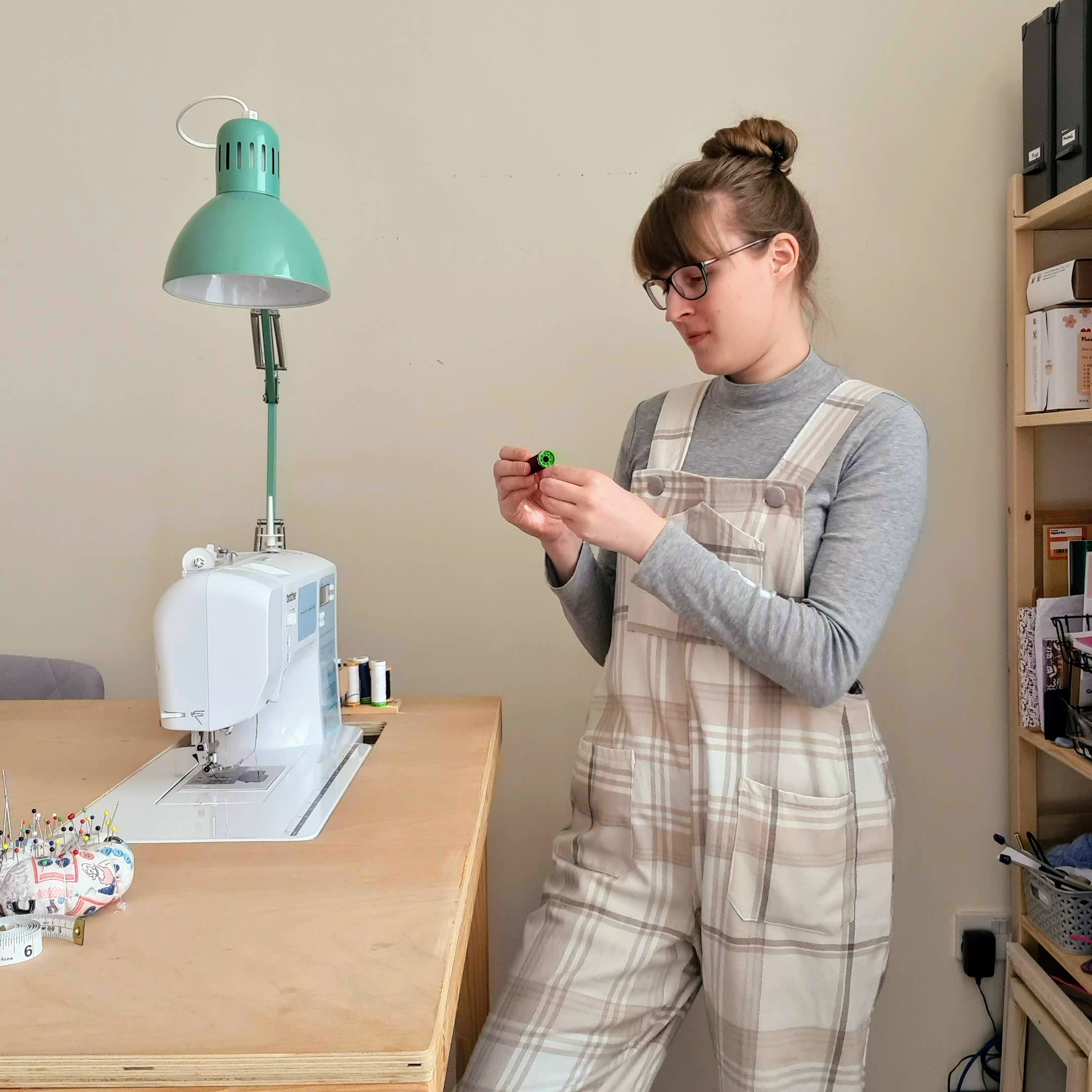 White female woman in twenties, wearing upcycled dungarees, standing next to her sewing machine.